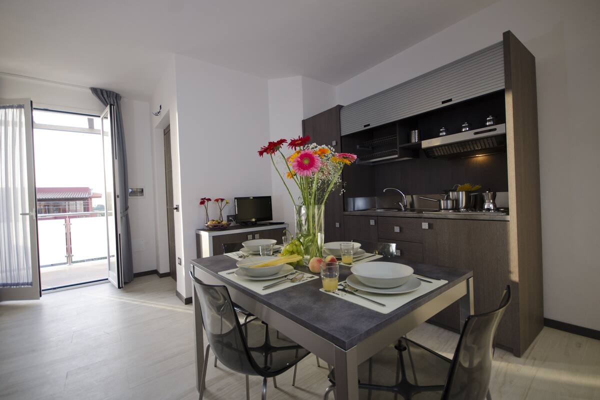 Una cocina moderna con armarios oscuros, una mesa de comedor lista para dos comensales y un florero con flores sobre la mesa.