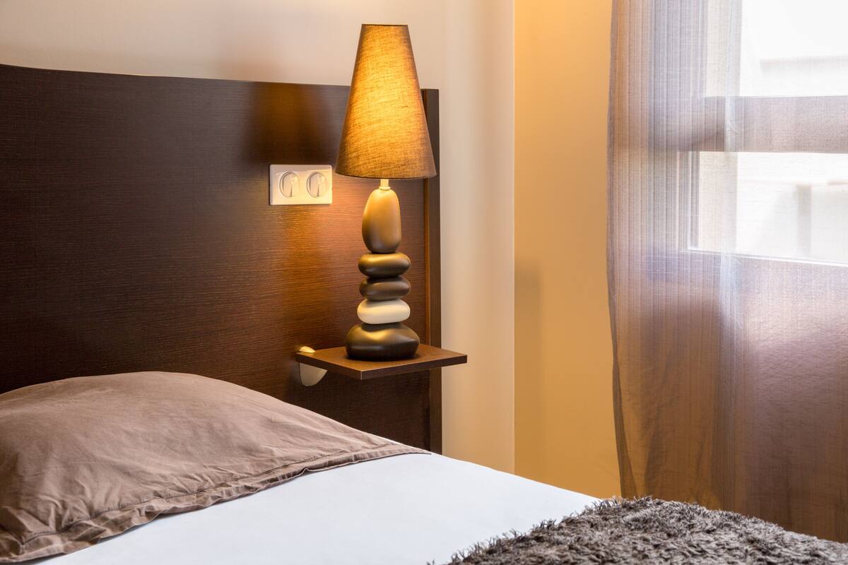 Ein ordentlich bezogenes Bett mit Kissen, eine Wandlampe und ein Fenster mit Vorhängen.