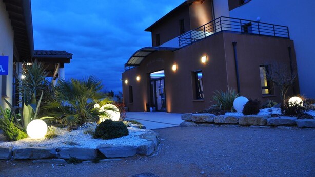A modern hotel entrance with a covered walkway, outdoor lighting, and landscaped garden areas. - The Originals City, Hôtel des Arts (Negrepelisse)
