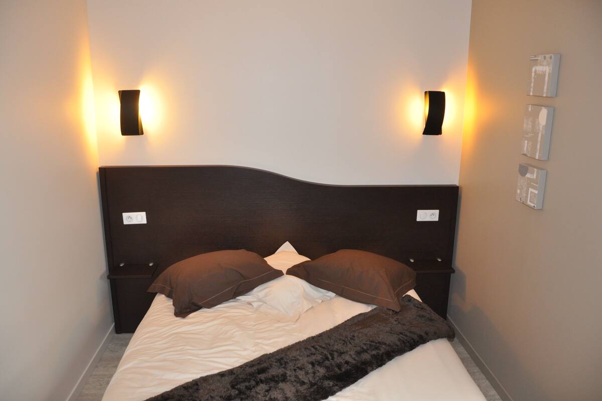 Ein ordentlich bezogenes Bett mit dunklem Kopfteil, zwei Wandleuchten und einem kleinen Nachttisch.