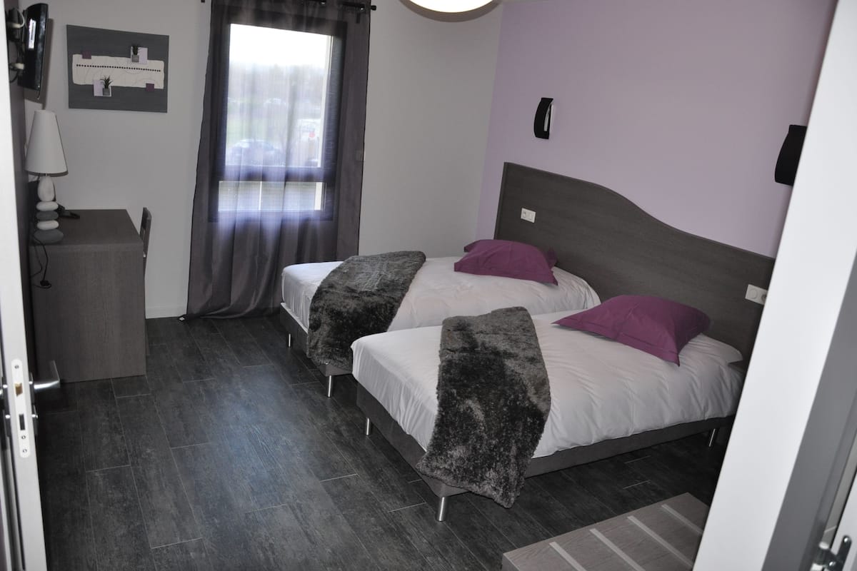 Ein Schlafzimmer mit zwei Betten, einem Schreibtisch mit Lampe, einem Fenster mit Vorhängen und einem an der Wand befestigten Telefon.