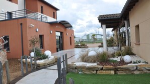 A modern building with a flat roof, a covered walkway, and a landscaped area with rocks and plants. - The Originals City, Hôtel des Arts (Negrepelisse)
