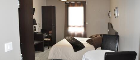 Une chambre à coucher avec un lit, un bureau et une chaise. Il y a une fenêtre avec des rideaux, un luminaire au plafond et une horloge murale.