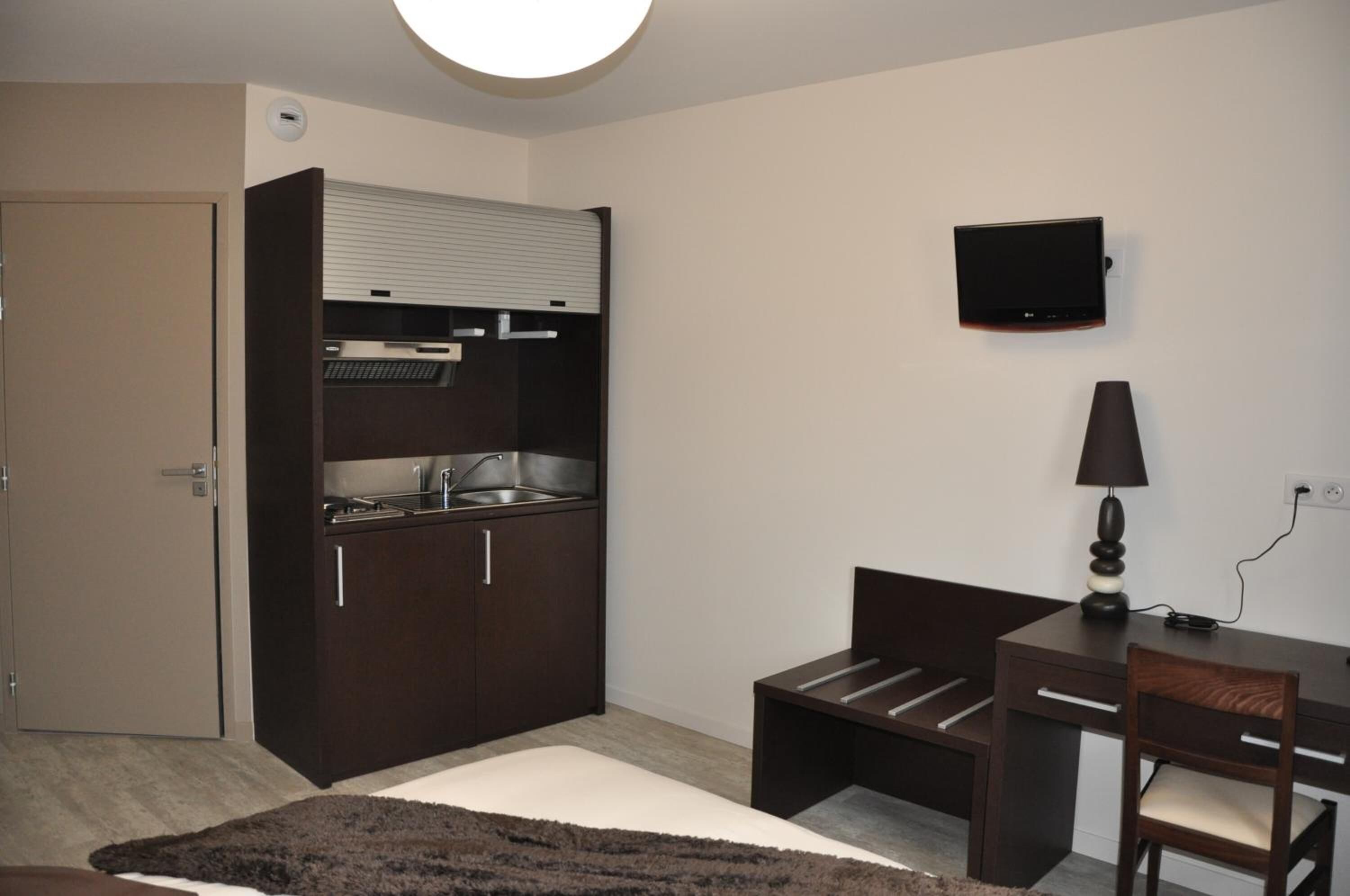 Une chambre compacte comprenant un coin cuisine, un téléviseur à écran plat, un bureau avec une chaise et une lampe.