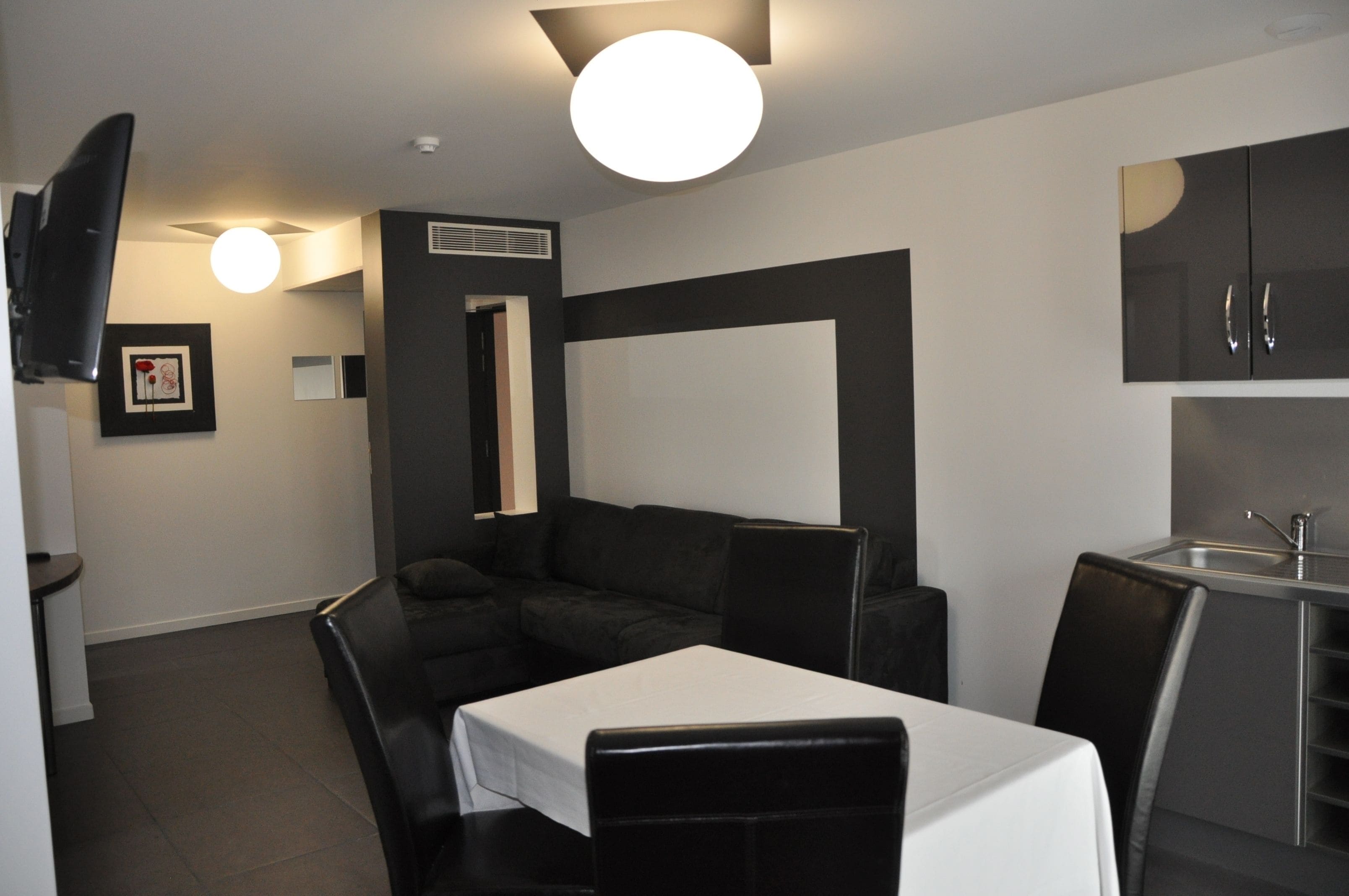 Habitación de hotel compacta con kitchenette, sofá, mesa de comedor y televisor montado en la pared.