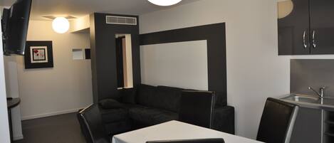 Habitación de hotel compacta con kitchenette, sofá, mesa de comedor y televisor montado en la pared.