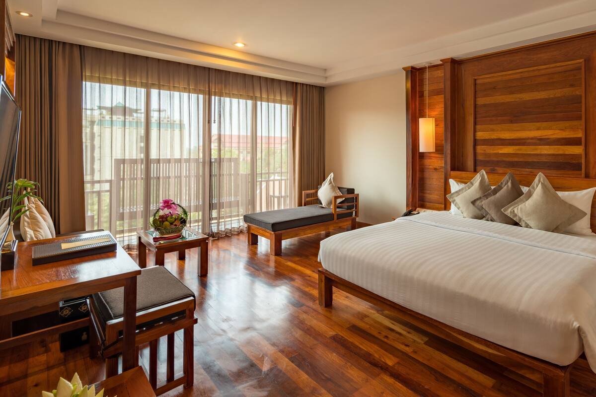 Habitación de hotel con una cama grande, suelo de madera, un escritorio con una silla, una mesita y un ventanal con cortinas.