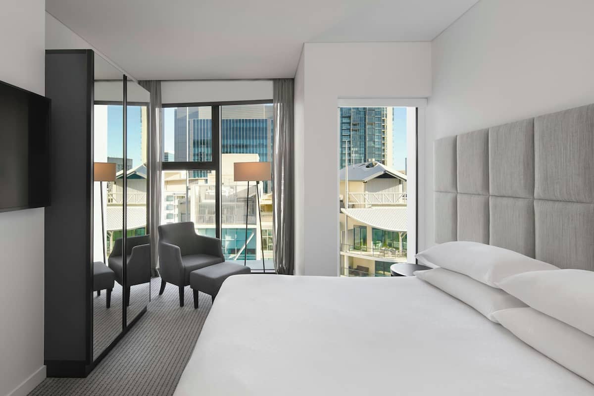 A modern hotel room with a large bed, a seating area with chairs, a TV, and a view of the cityscape through large windows.