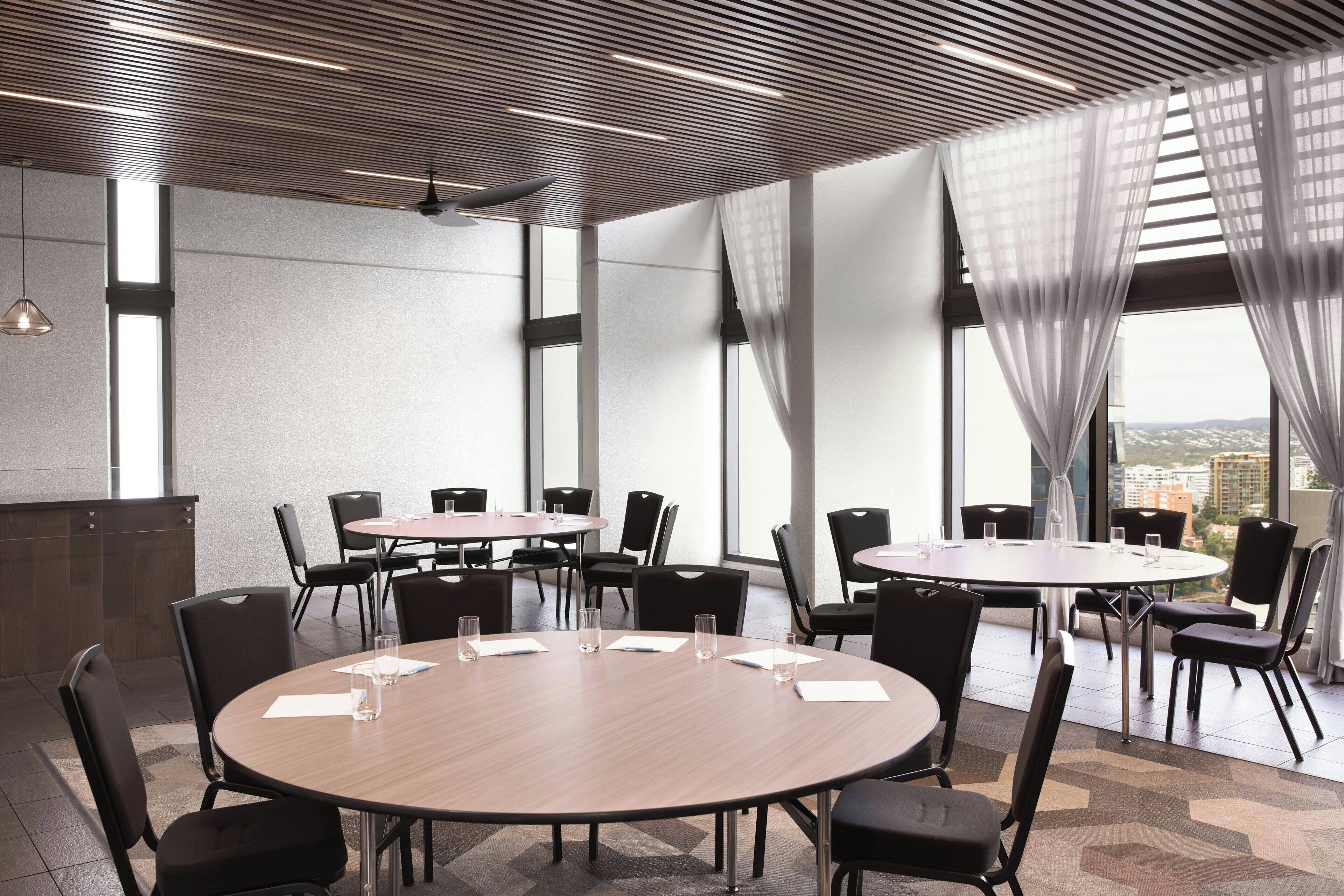 A conference room with round tables, black chairs, and large windows with sheer curtains.