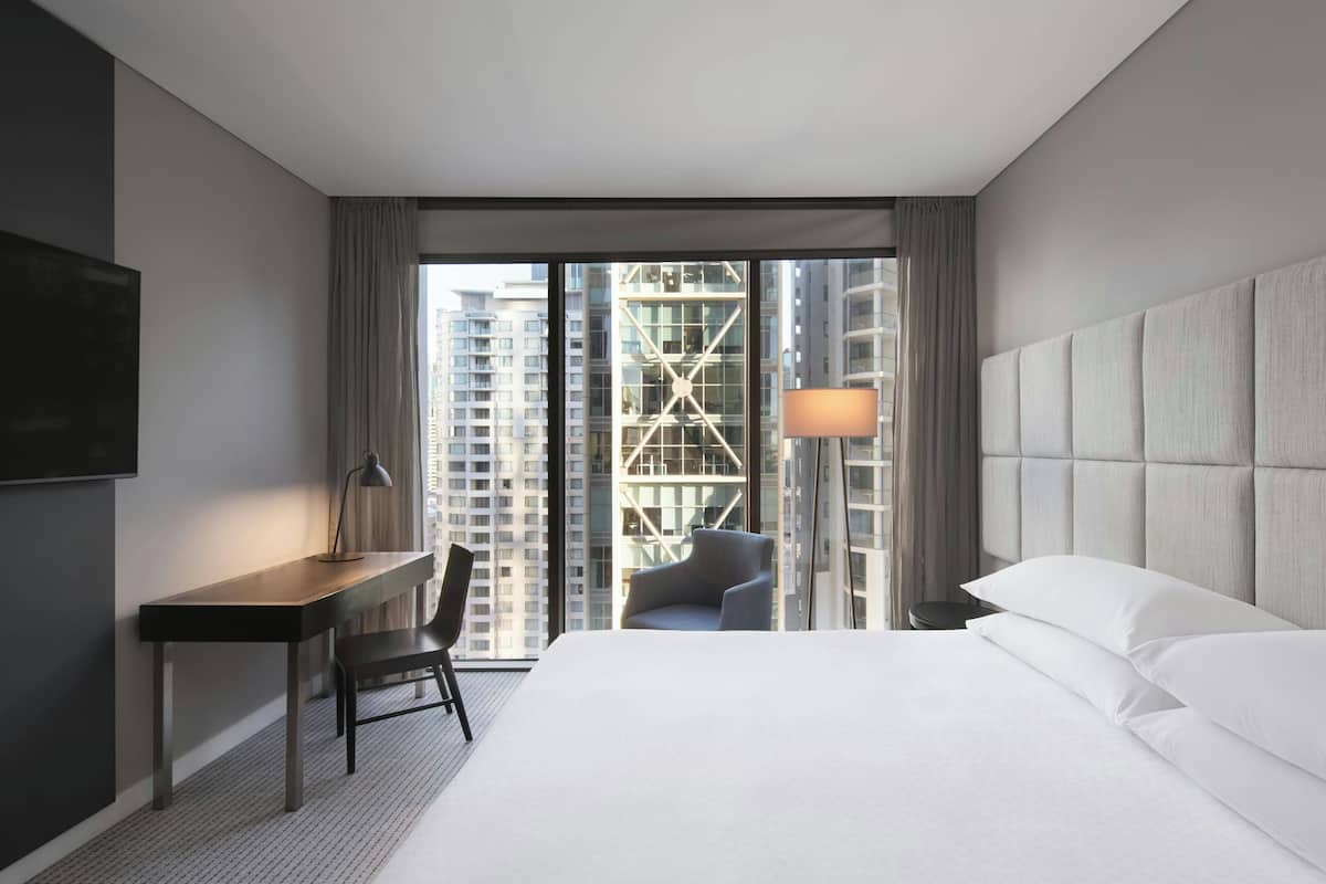 A hotel room with a bed, a desk, a chair, a lamp, and a view of tall buildings through the window.