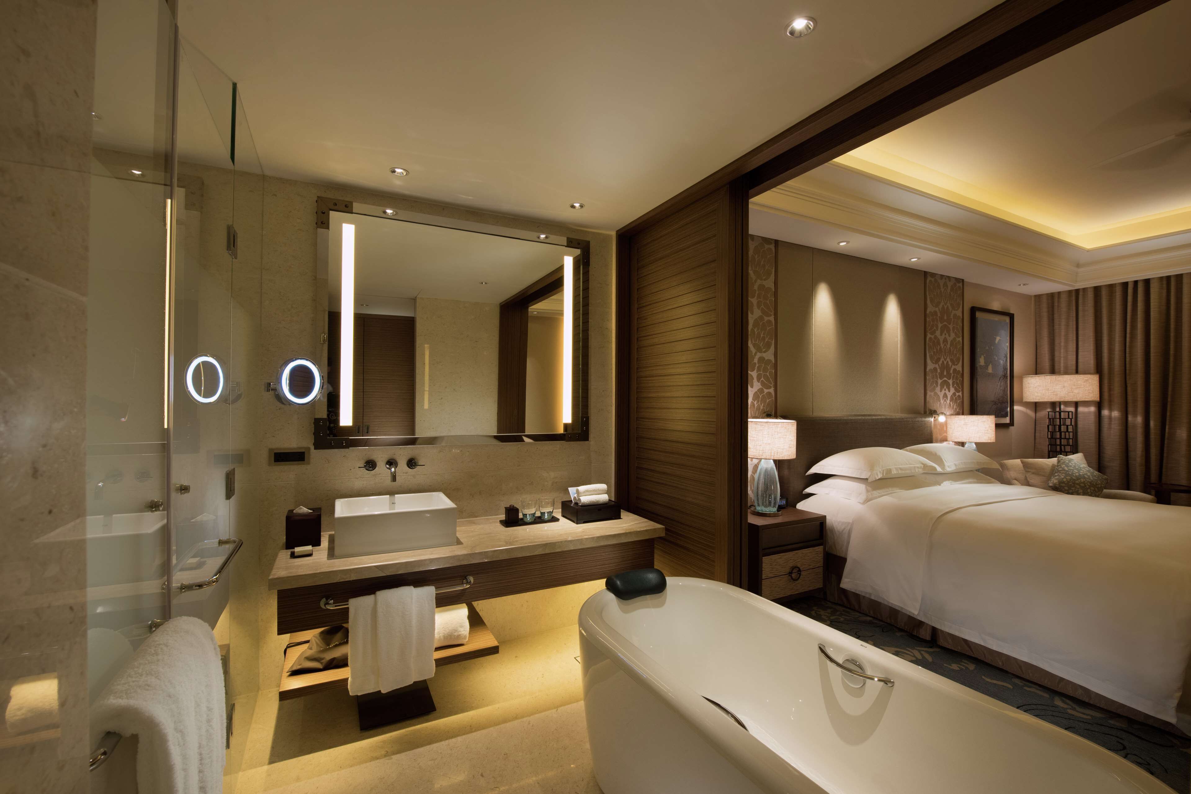 A modern hotel room with a glass shower, a large mirror, a bathtub, a bed with white linens, and a bedside table with a lamp.