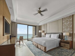 A spacious hotel room with a large bed, a ceiling fan, a balcony with a view of the sea, and a flat-screen TV.