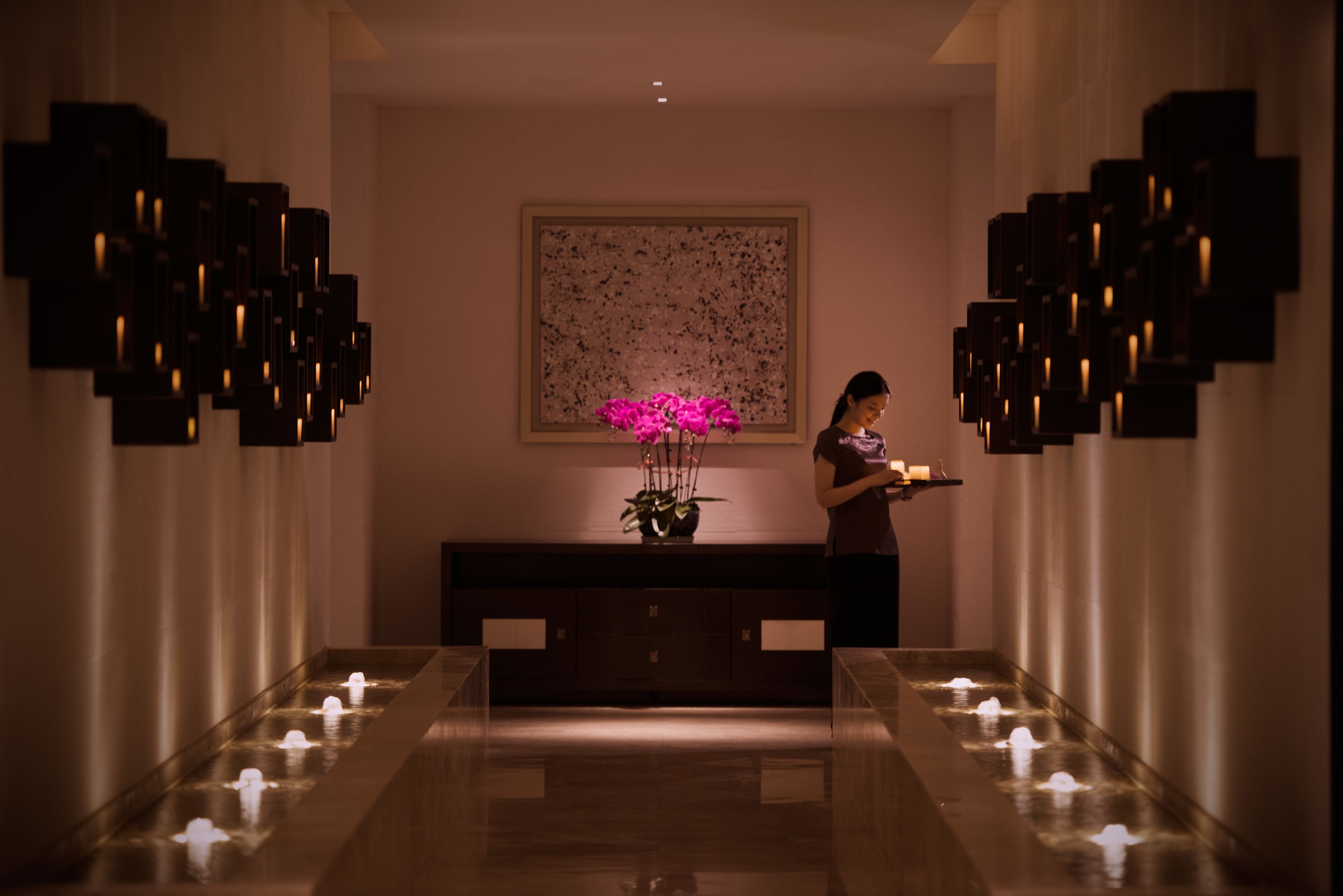 A hallway with a long bench, a wall-mounted sculpture, and a potted plant with pink flowers.
