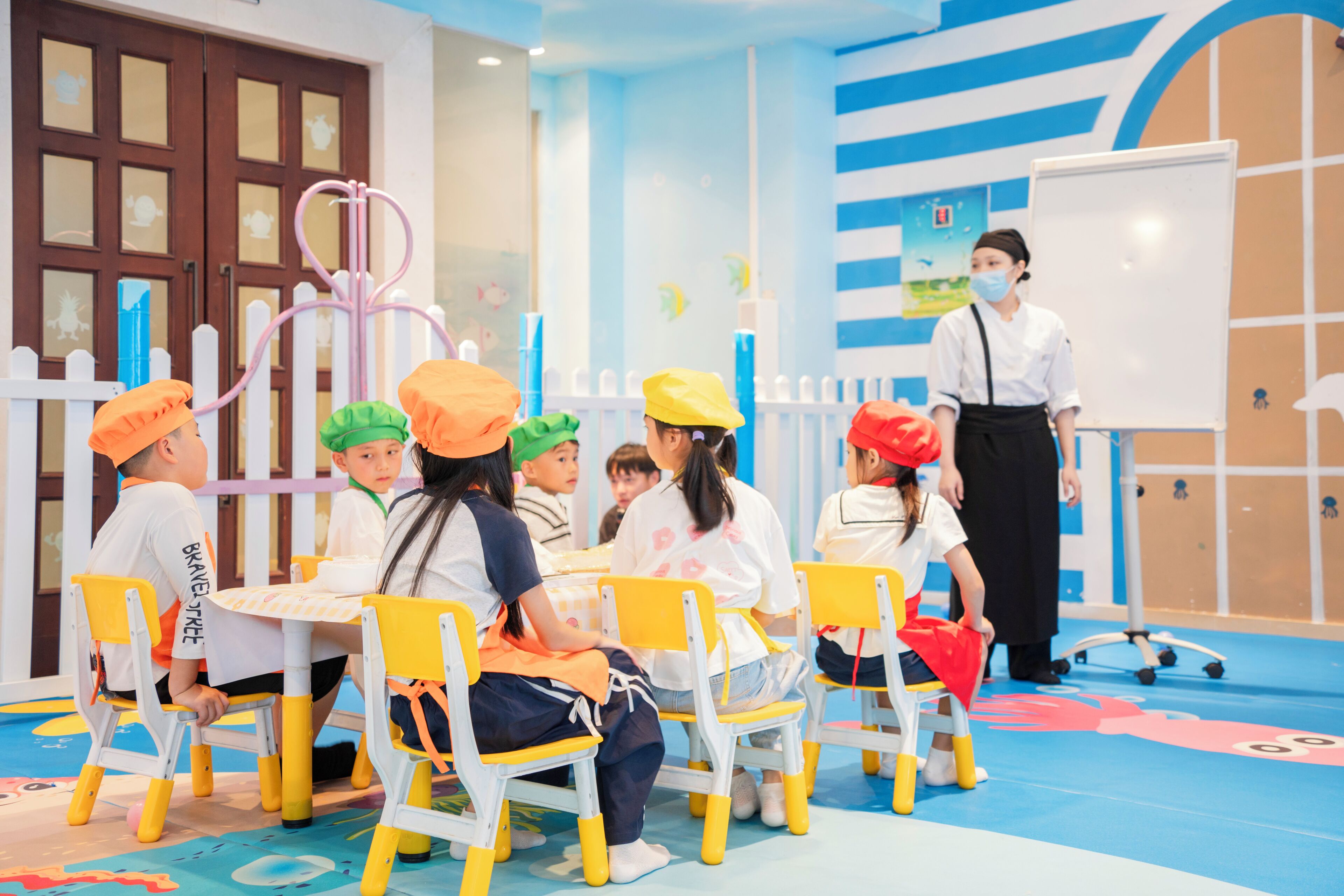 Een groep kinderen in een klaslokaal met een leraar, gele stoelen en een whiteboard.