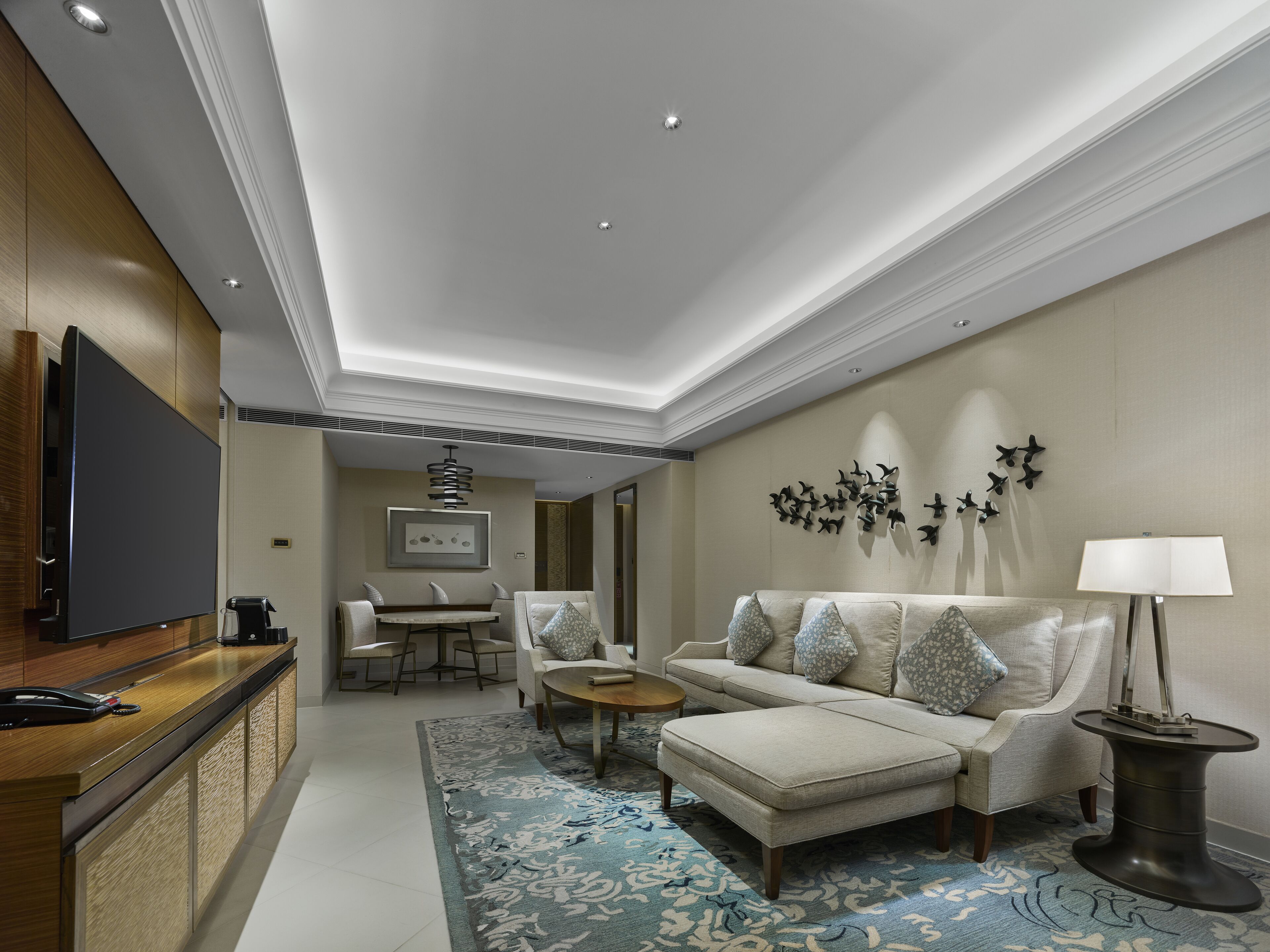 A modern hotel room with a flat-screen TV, a wooden desk, a sofa, a coffee table, and a patterned rug.