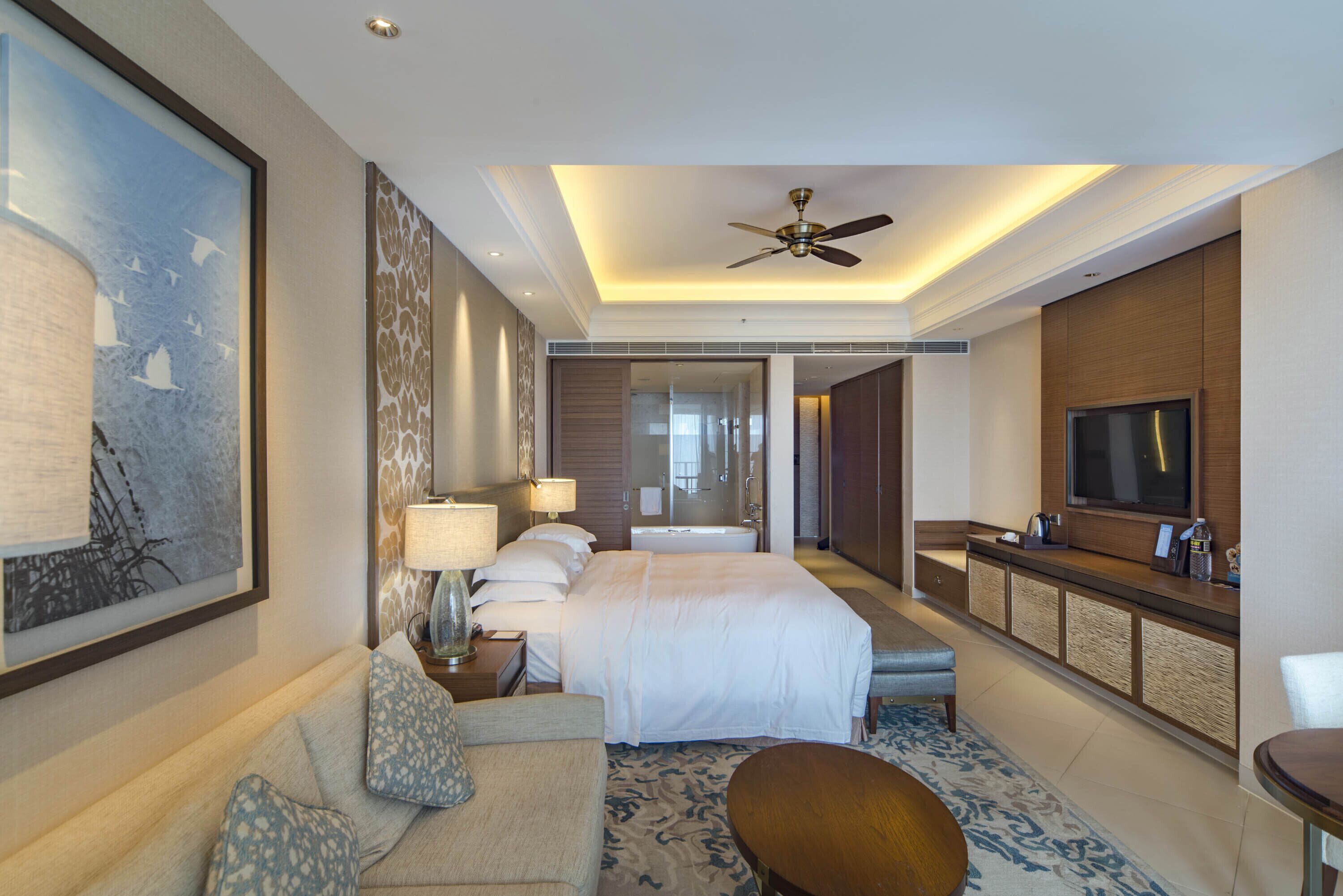 A hotel room with a large bed, a desk with a TV, a ceiling fan, and a large window.