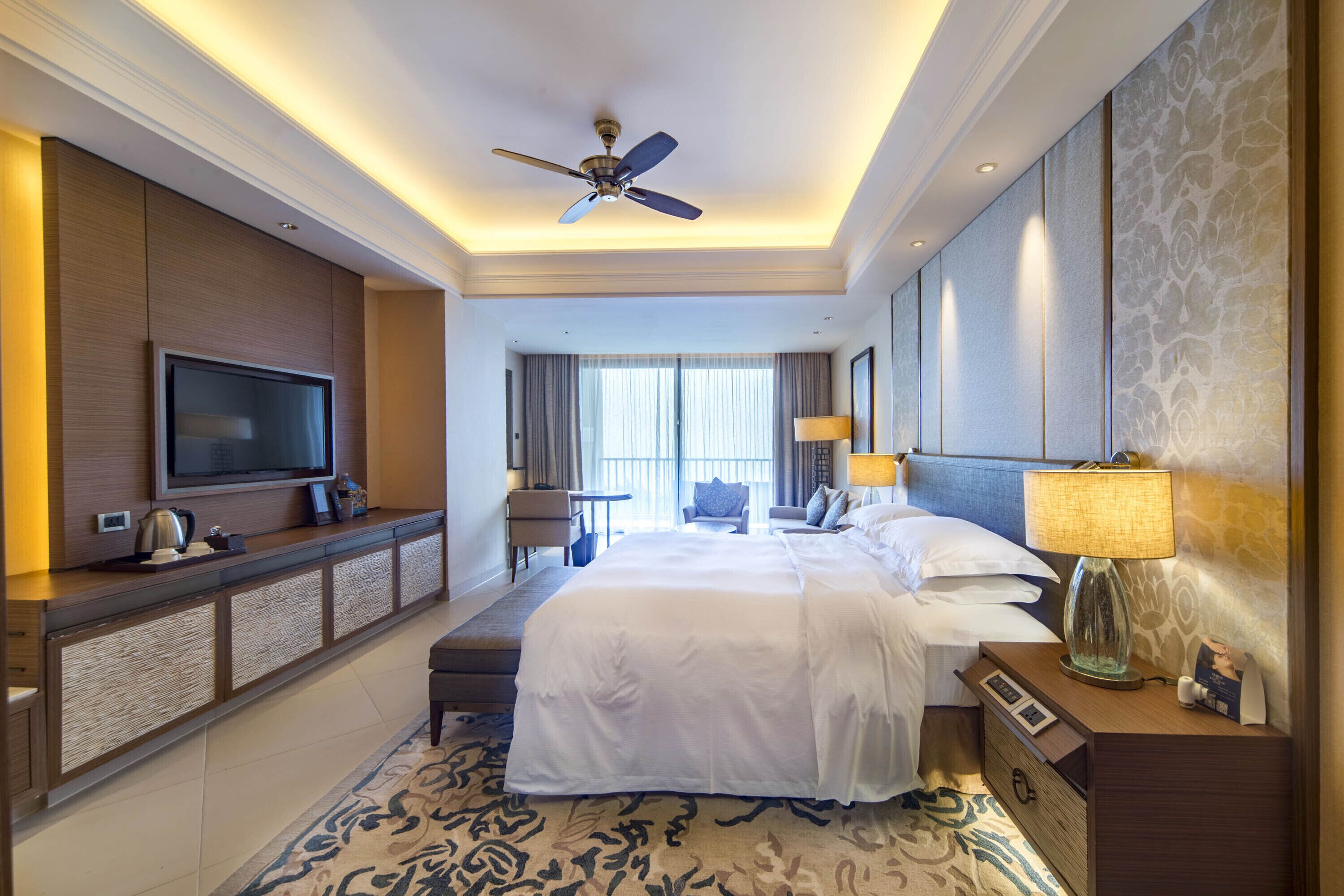 A hotel room with a large bed, a TV on a wooden console, a ceiling fan, a desk with a lamp, and a view of the outdoors through large windows.
