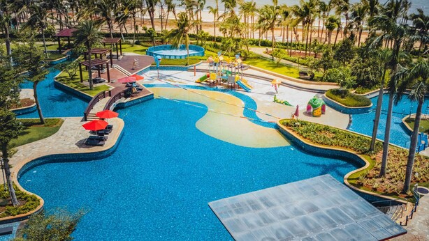 A large, winding pool with a view of the ocean, surrounded by palm trees and a beach. - Hilton Haikou Meilan (Haikou)