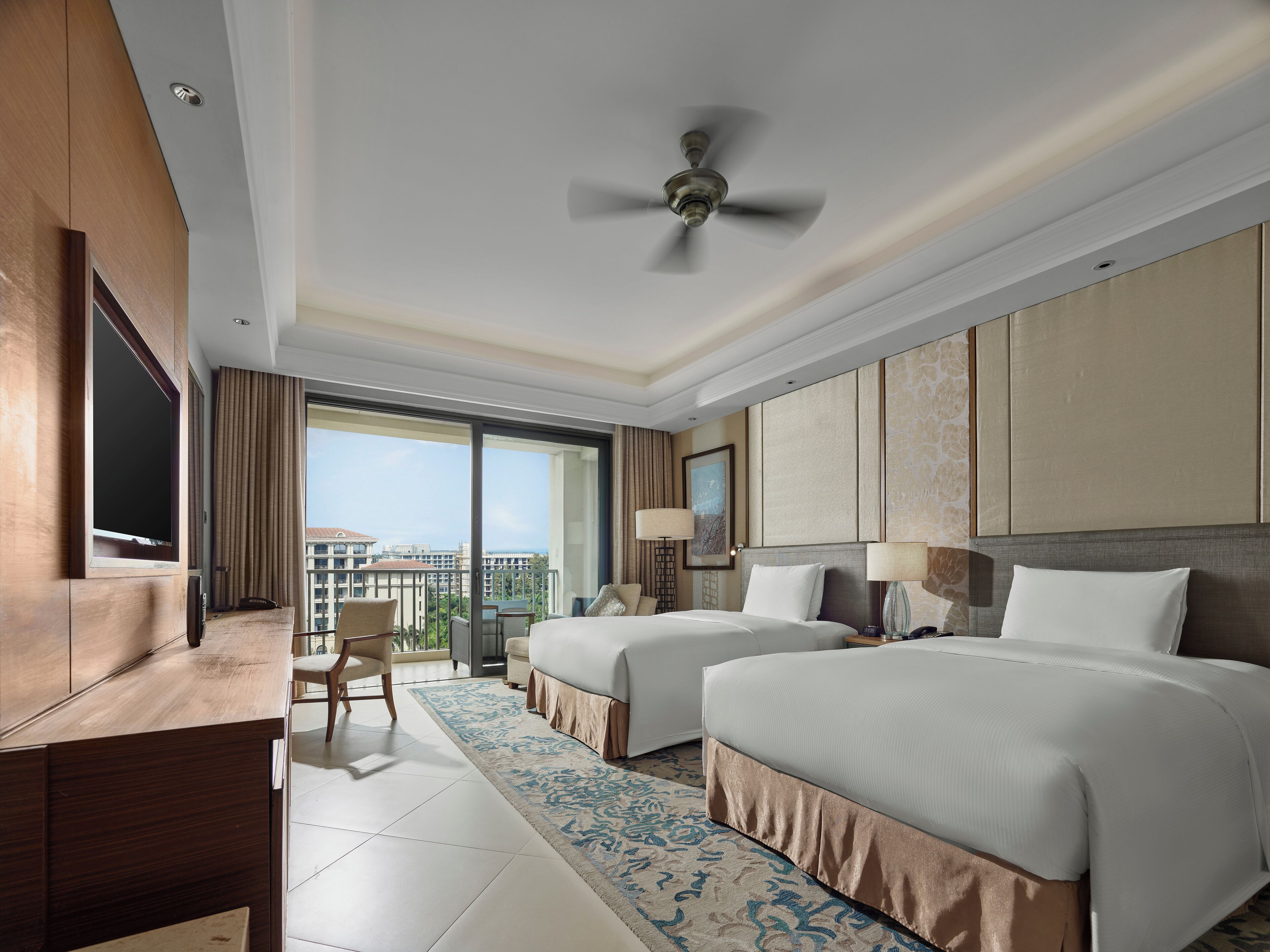 A hotel room with two beds, a TV, a desk, and a balcony with a view.