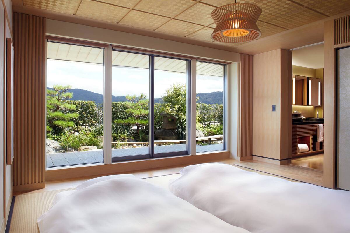 A bedroom with a large window offering a view of a garden and mountains.