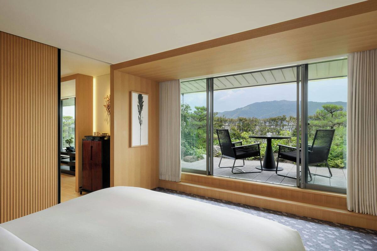 A hotel room with a large bed, a view of the outdoors through a sliding glass door, and a small table with chairs outside.