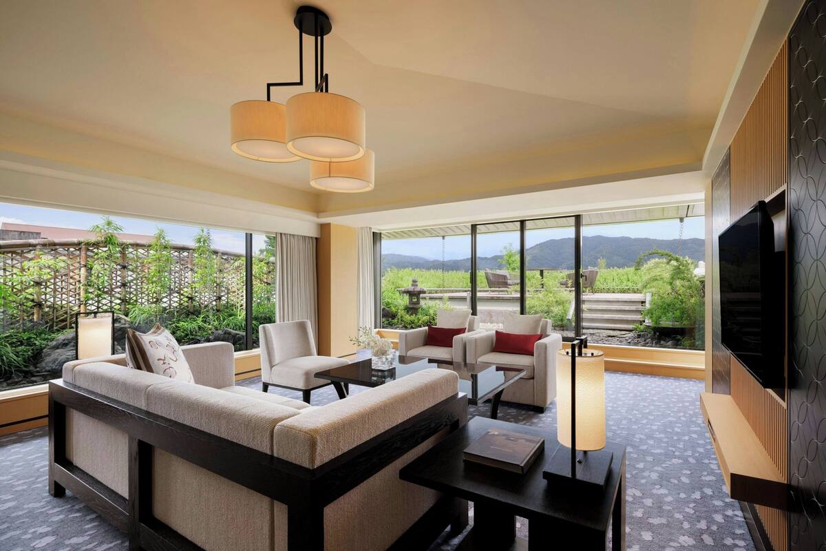 A modern hotel room with a large sofa, two armchairs, a coffee table, and a TV. The room has large windows offering a view of greenery and mountains.
