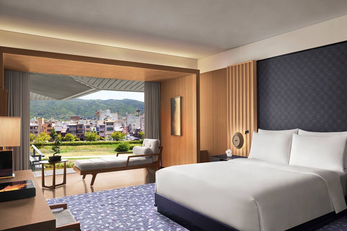 A modern hotel room with a large bed, a seating area with a view, and a balcony with a cityscape view.