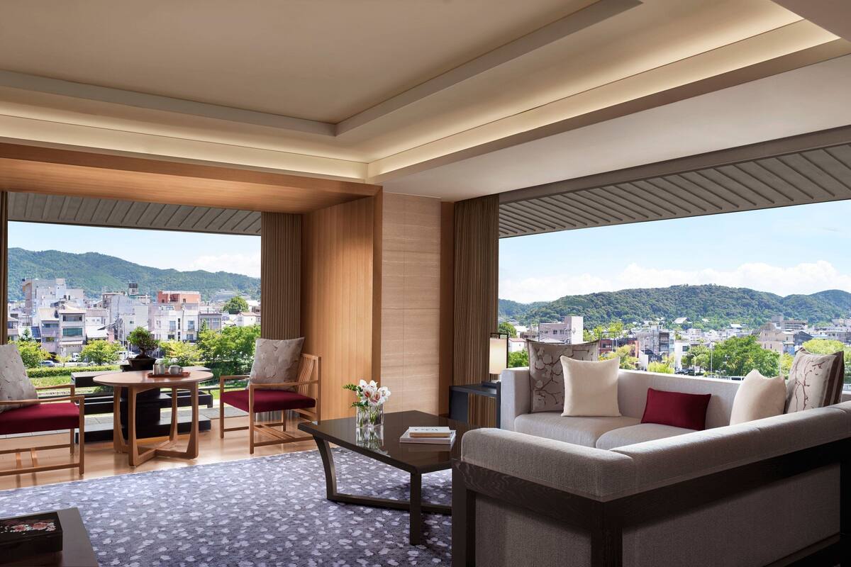 A modern hotel room with a large window offering a city view, a sofa, a dining table, and a chair.