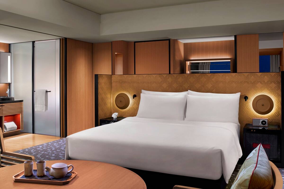 A hotel room with a large bed, a bedside table with a radio, a wooden headboard, and a small seating area with a tray of tea cups.