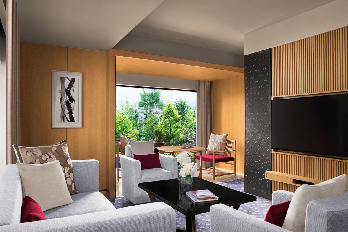 A modern living room with a sofa set, a coffee table, and a flat-screen TV. There is a large window offering a view of greenery outside.