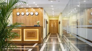 Zona de recepción de un hotel, con piso de mármol, mostrador de madera, relojes en la pared y una planta en maceta.