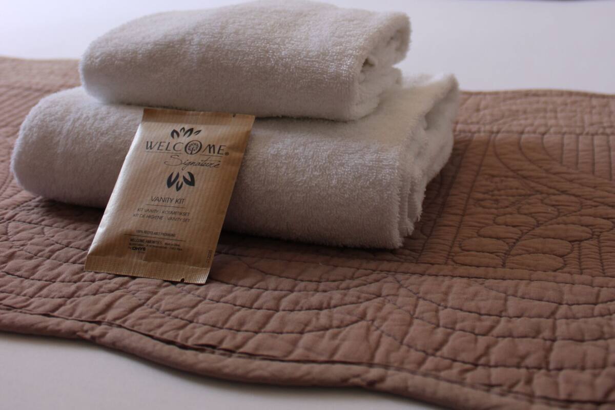 Un lit bien fait, avec une couverture marron, une pile de serviettes pliées et un kit de bienvenue posé sur le lit.