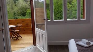 A room with a bed, a wooden table, and a view of greenery through large windows.