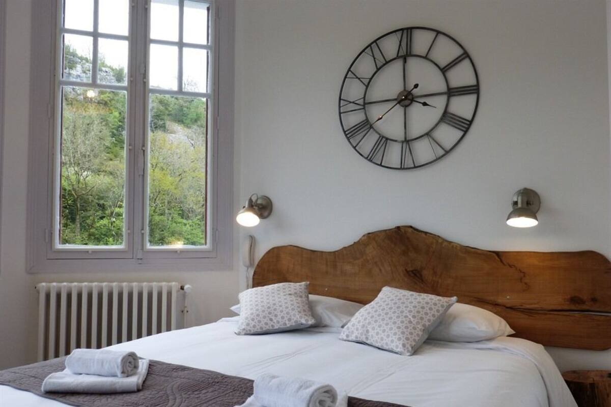 Un lit bien fait, avec une tête de lit en bois, une grande horloge murale et deux lampes fixées au mur.