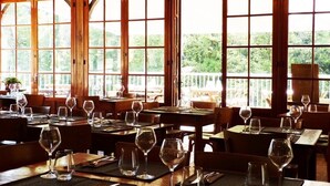Ein Restaurant mit Holztischen und -stühlen, großen Fenstern mit Blick auf Grünflächen und freundlicher Beleuchtung.