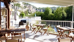 Eine Außenpatio mit Holztischen und -stühlen, einem weißen Geländer und Blick auf einen Fluss und Berge.