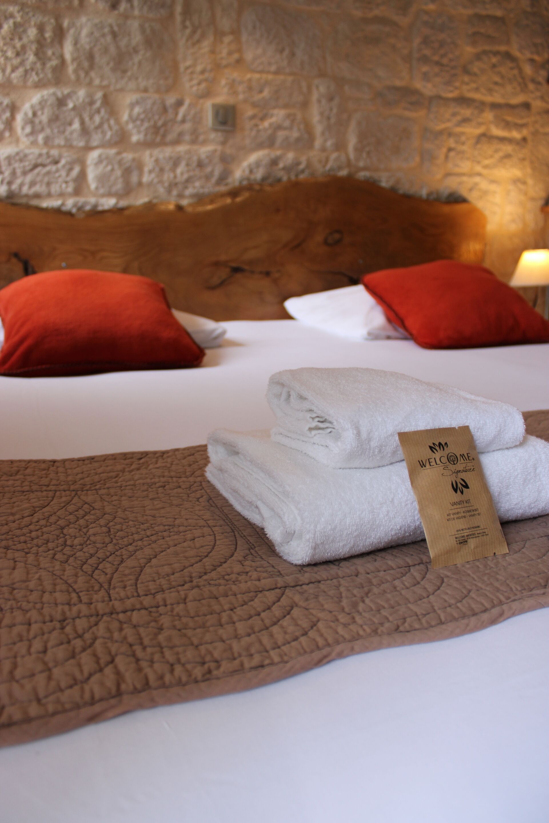 A neatly made bed with white linens and two red pillows, a folded towel, and a welcome card on the bed.