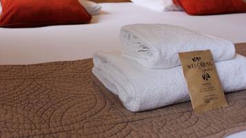 A neatly made bed with white linens and two red pillows, a folded towel, and a welcome card on the bed.