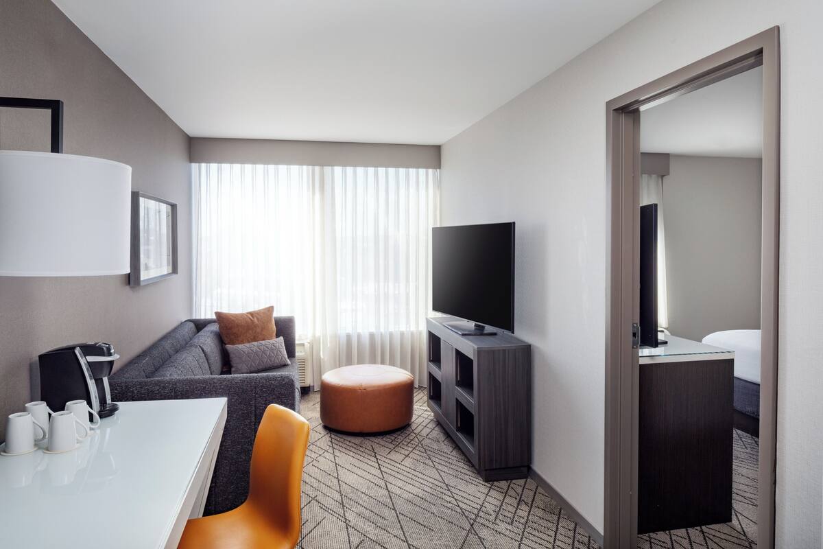 A modern hotel room with a sofa, ottoman, TV, and a dining area with a table and chairs.