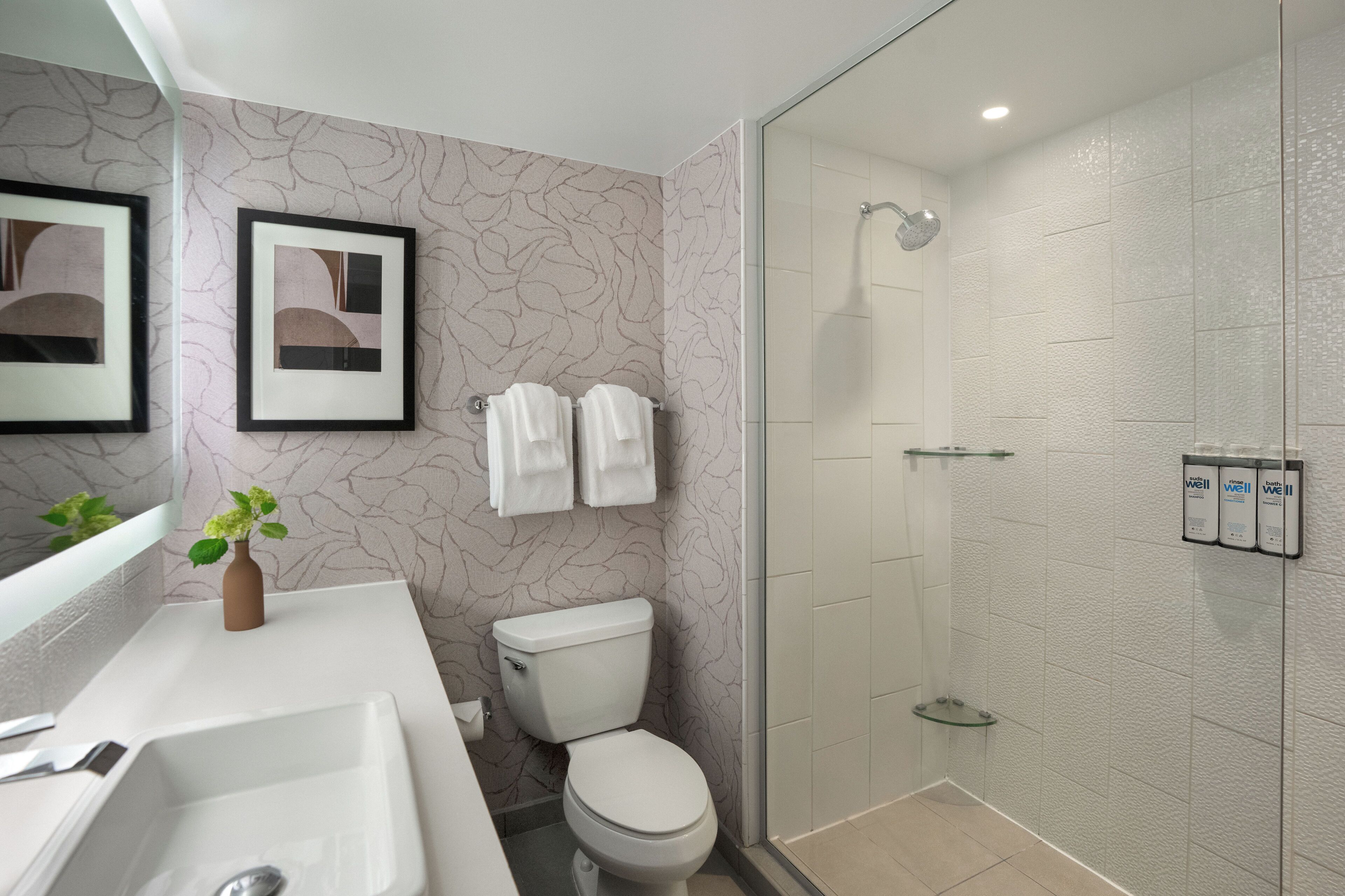 A modern bathroom with a white sink, toilet, and shower. There are framed pictures on the wall and a vase with flowers on the counter.