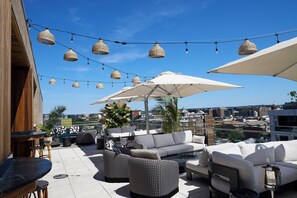 An outdoor seating area with multiple couches, tables, and umbrellas, overlooking a cityscape.