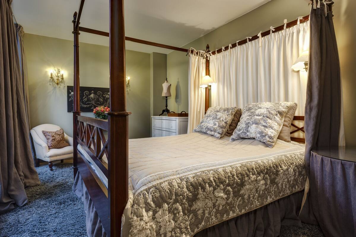 Una cama con dosel con un edredón floral, una mesita de noche con una lámpara, una silla y un cuadro en la pared.