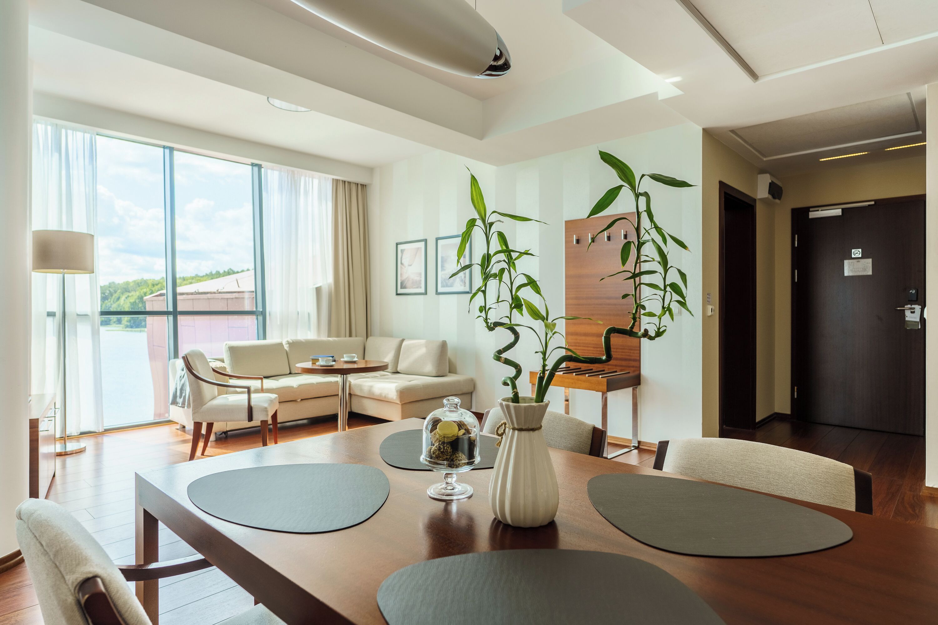 A modern hotel room with a dining area, a sofa set, a large window, and a potted plant.