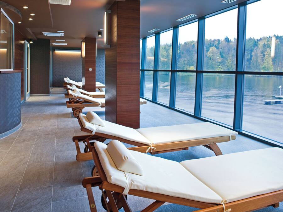 Zona de spa con tumbonas ordenadas a lo largo de un pasillo, amplias ventanas con vistas a un lago y un bosque.