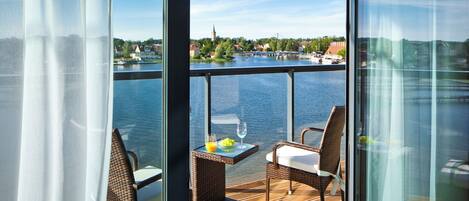 A balcony with wicker furniture, a table with a glass of wine and fruit, and a view of a river and buildings.