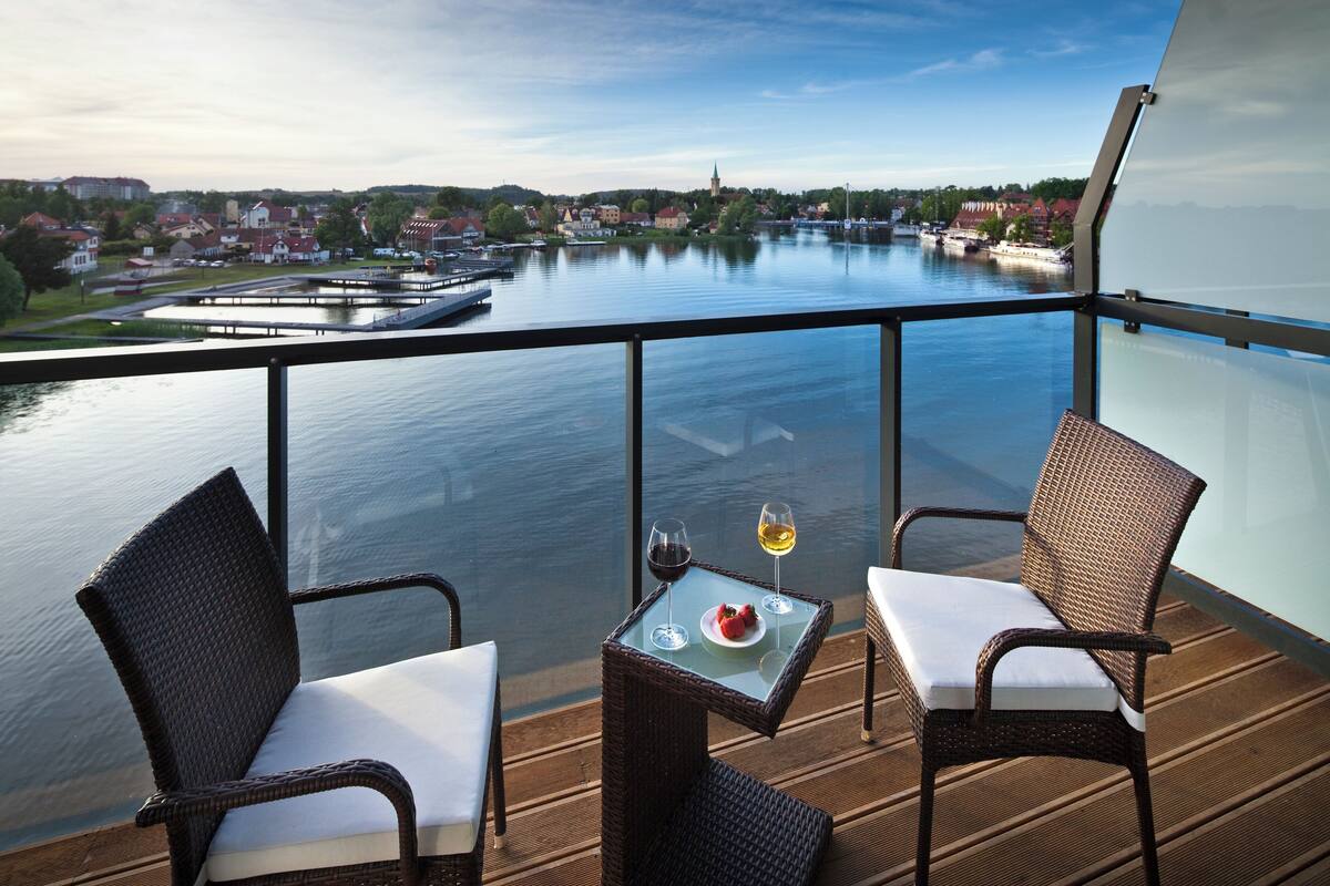 Een balkon met rieten stoelen en een tafel, met uitzicht op een rustige rivier met gebouwen op de achtergrond.