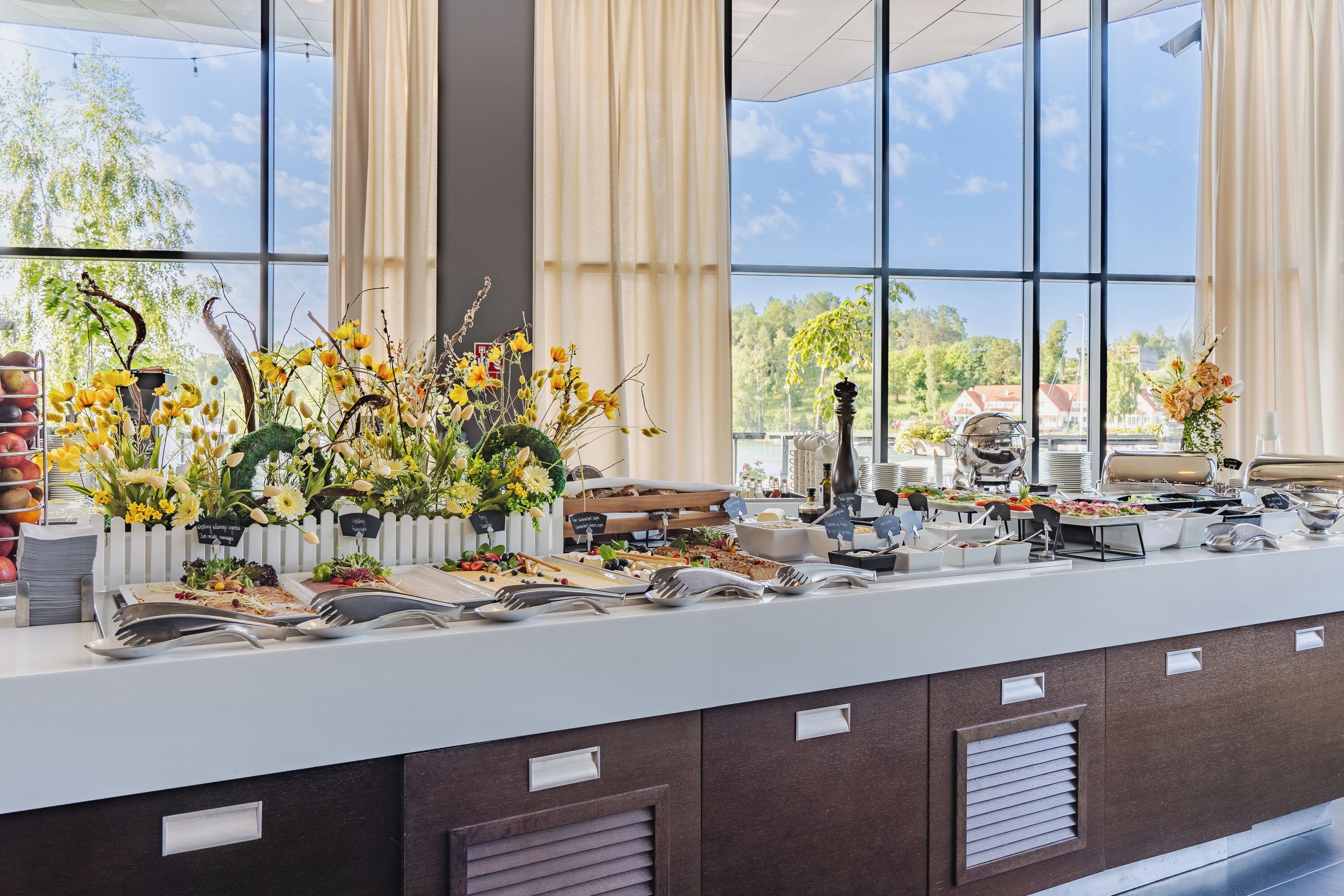 A buffet setup with various food items and utensils, a white fence, and a view of greenery and buildings through large windows.