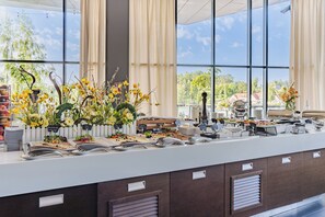 A buffet setup with various food items and utensils, a white fence, and a view of greenery and buildings through large windows. - Hotel Mikołajki Leisure & SPA - Destigo Hotels (Mikolajki)