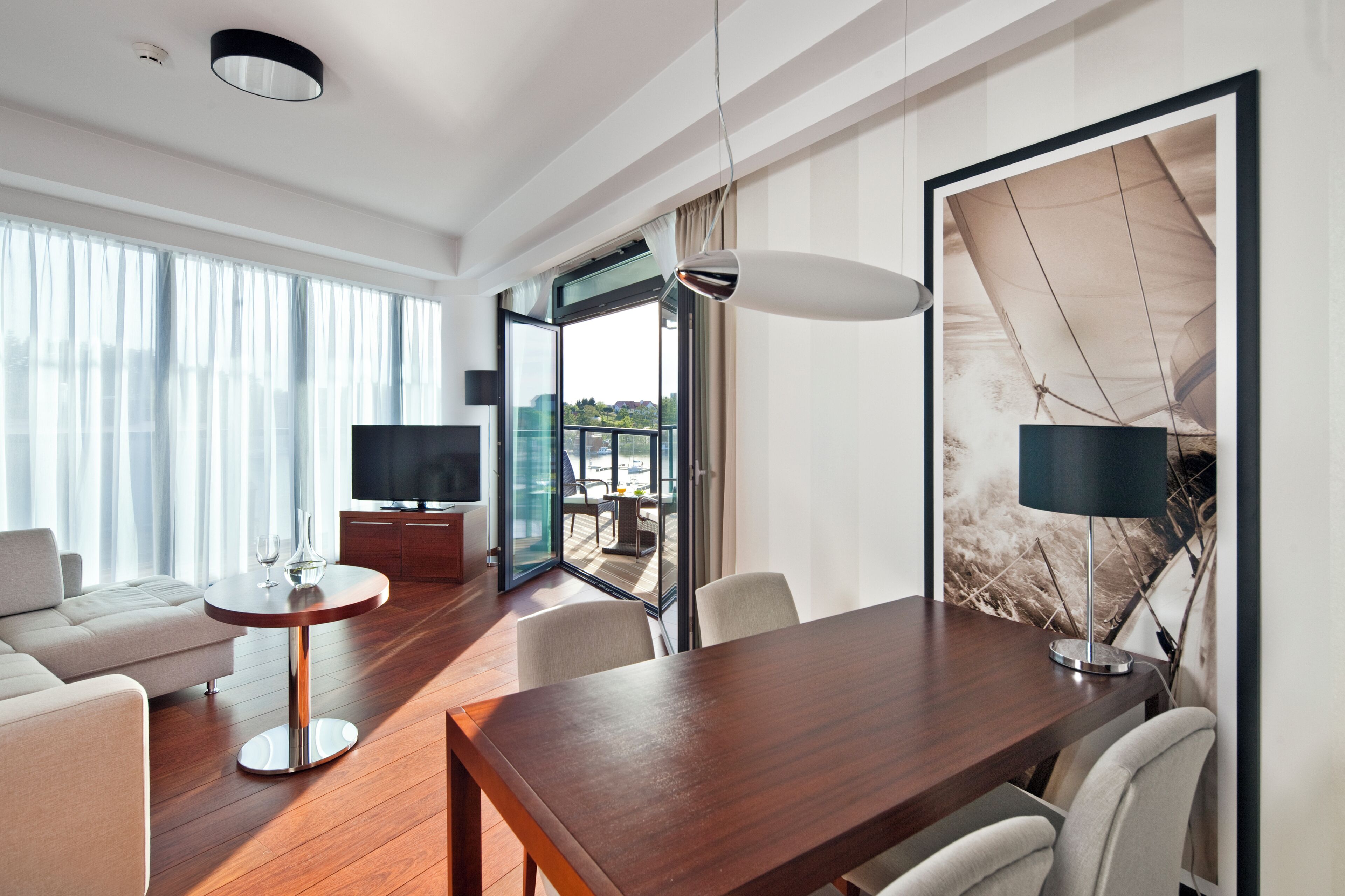 A modern hotel room with a wooden floor, a dining table, a sofa, a TV, and a large window with curtains.