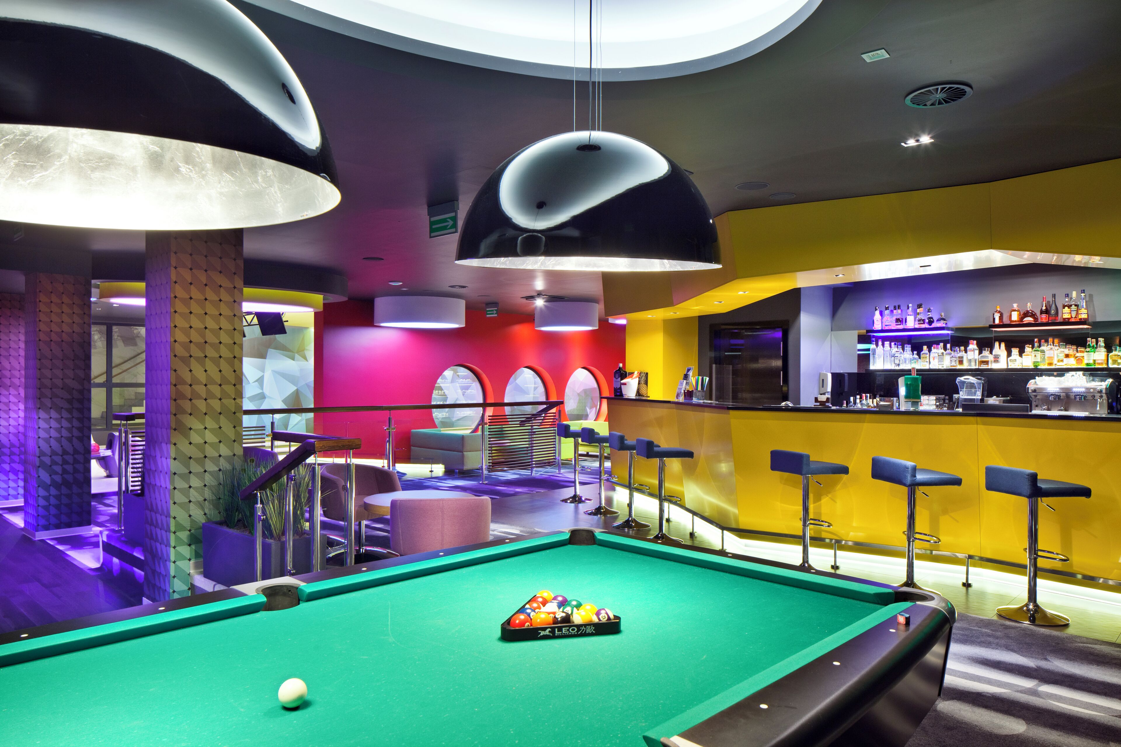 A pool table with a billiard ball rack in a brightly lit bar area with a yellow wall and a bar counter.